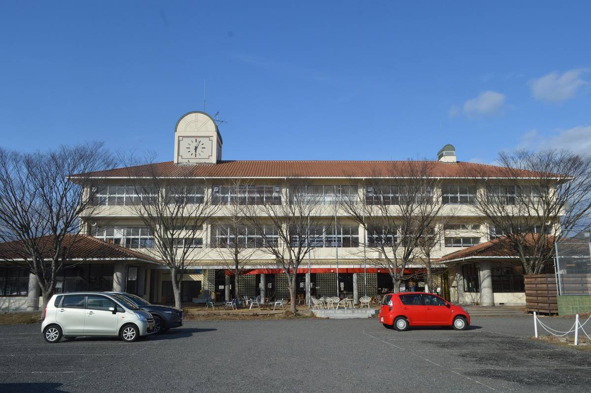 冬の晴れた日の、3階建ての校舎。屋根の中央に時計塔がある。1階の前にテラス席のいすと机が並び、手前は駐車場で車が3台止まっている。人は写っていない。