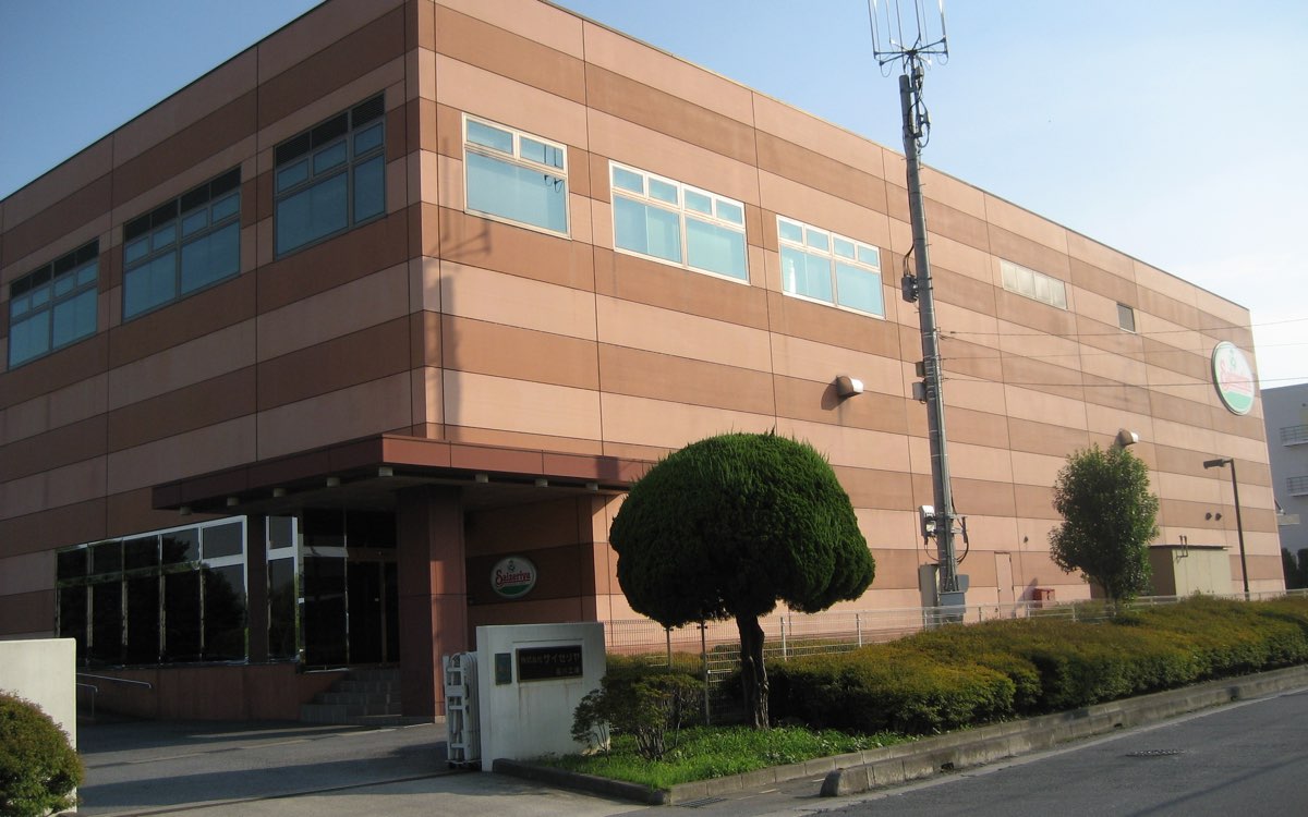 晴れた夕方の、2階建ての大きな建物の外観。薄茶色と茶色の横じまの外壁に窓が並び、入口の前に丸く刈った木と社名の門柱がある。人は写っていない。